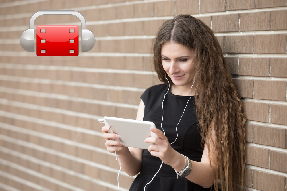 Una mujer ve la televisión en una tableta mientras escucha por unos auriculares y aparece el logo de AudescMobile