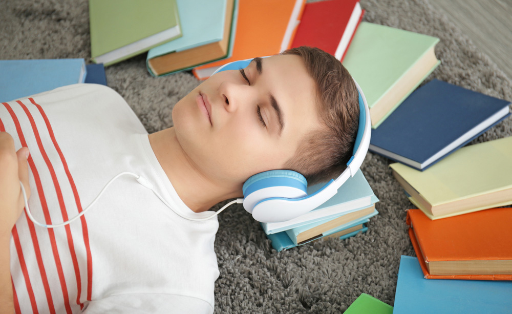 Un joven escucha un libro en formato digital tumbado y rodeado de libros en formato físico