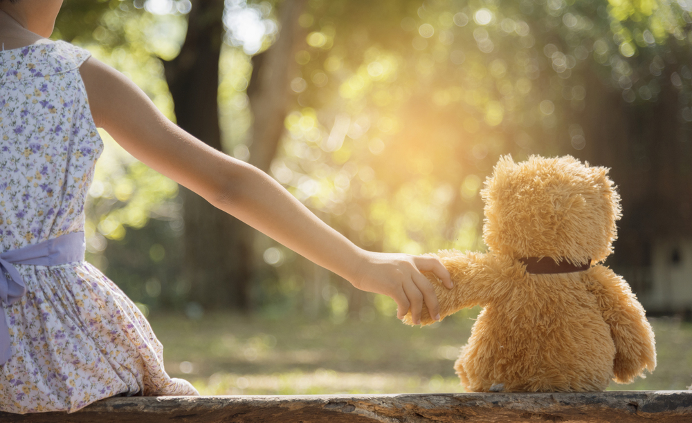 Una ni&ntilde;a sentada en el tronco de un &aacute;rbol sujeta la mano de un oso de peluche