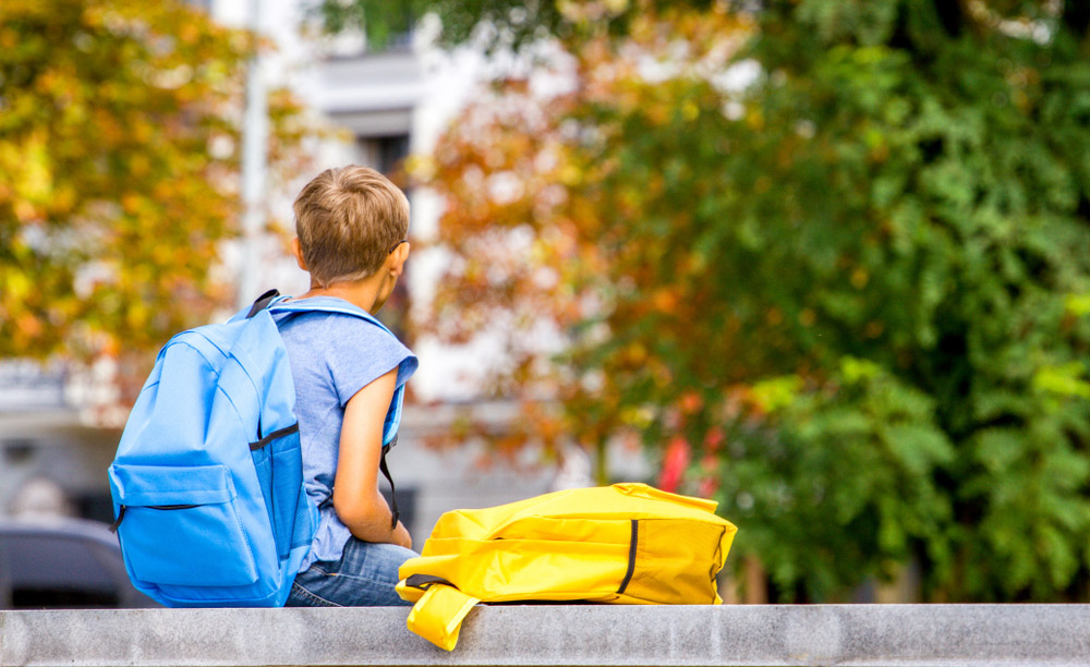 Un niño espera sentado en el exterior de un colegio con una mochila azul a la espalda