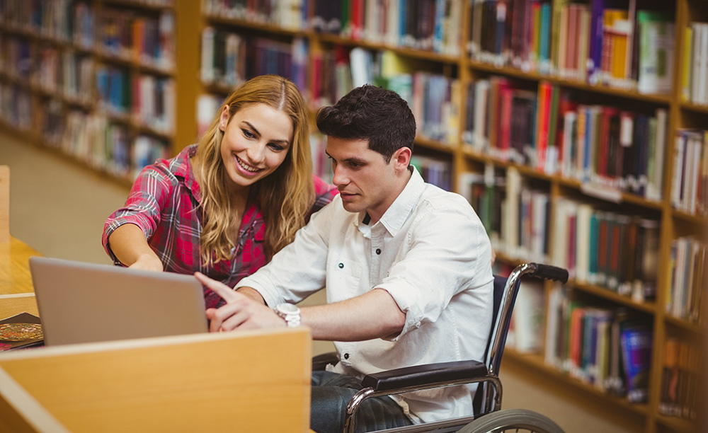 Un joven en silla de ruedas junto a otra joven estudian en una biblioteca con un ordenador