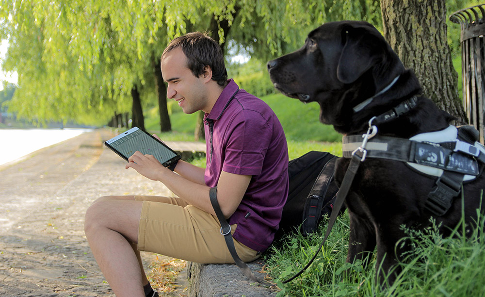 Un joven con discapacidad visual utiliza una tableta de Feelif sentado en un parque con su perro al lado
