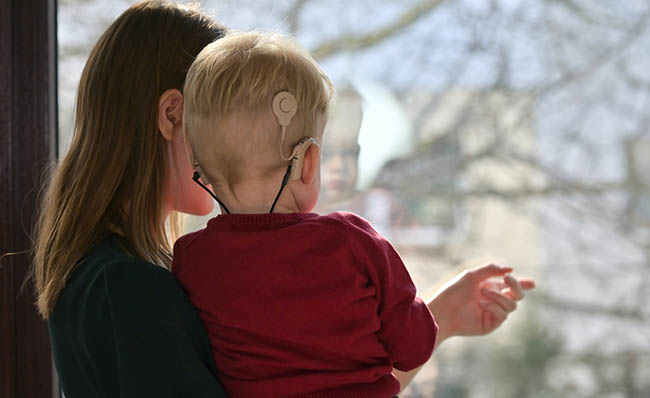 Un niño con dos implantes cocleares mira a través de una ventana en brazos de su madre