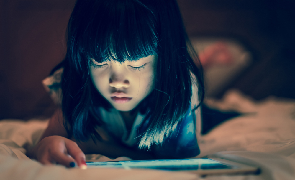 Una niña utiliza una tableta a oscuras tumbada sobre una cama