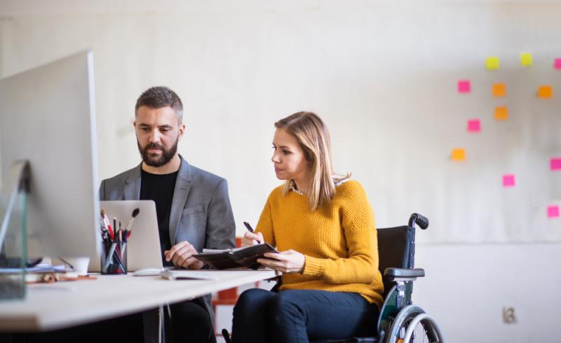 Una mujer en silla de ruedas en una oficina delante de un ordenador y junto a un compañero