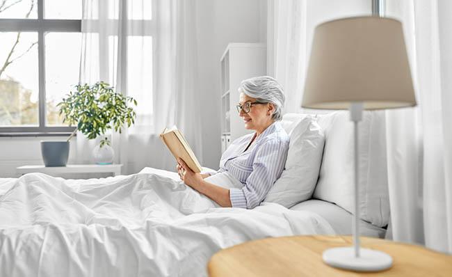 Una mujer mayor lee en su cama con un dispositivo Alexa al fondo