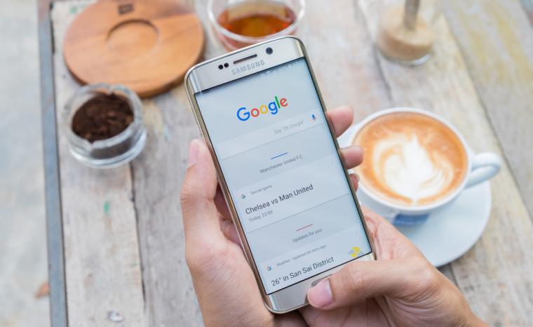 Una personas sujeta un tel&eacute;fono m&oacute;vil encima de una mesa en la que hay un caf&eacute;