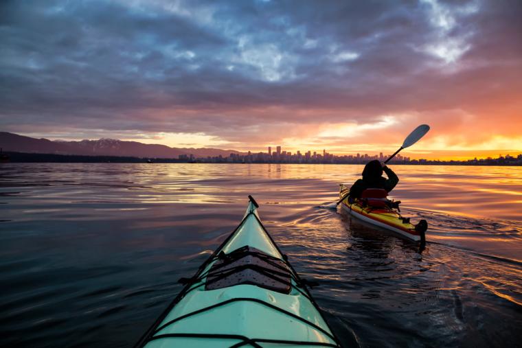 Un kayak en primer plano y una persona montando en otro kayak, con una ciudad de fondo al atardecer