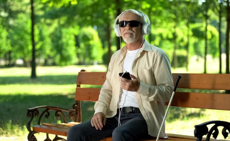 Una hombre con discapacidad visual utiliza su tel&eacute;fono m&oacute;vil sentado en un banco
