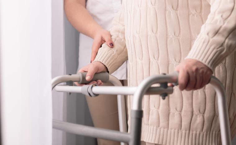 Una persona mayor utiliza un andador acompa&ntilde;ado por un cuidador
