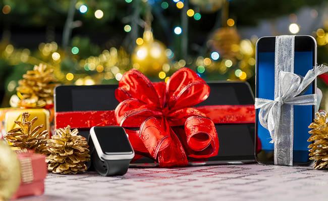 Imagen de dispositivos tecnol&oacute;gicos envueltos y colocados delante de un &aacute;rbol de Navidad