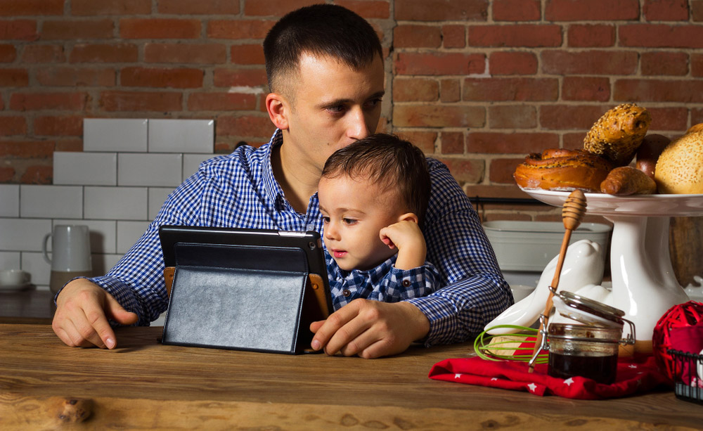 Un padre trabaja desde su casa usando una tableta y con su niño pequeño en brazos
