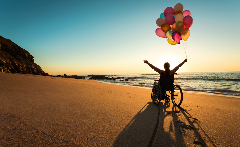 Una persona en silla de ruedas disfruta de la playa con globos de muchos colores cogidos de una mano