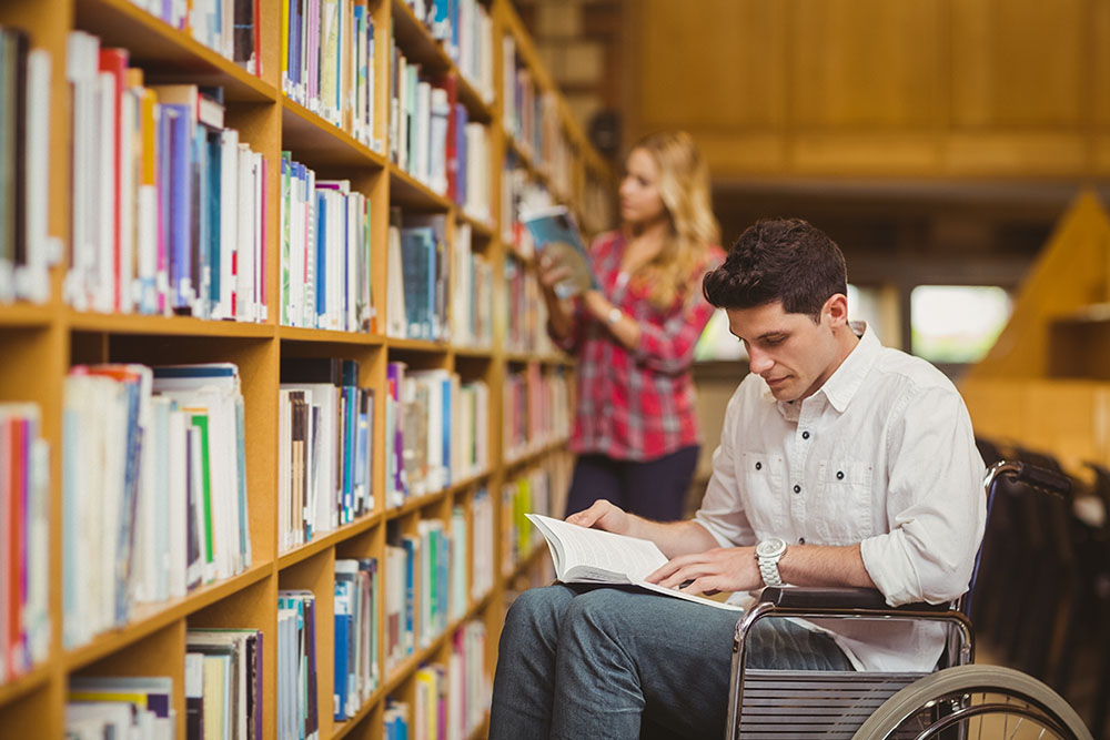 Un joven en silla de ruedas consulta un libro en la biblioteca de una universidad