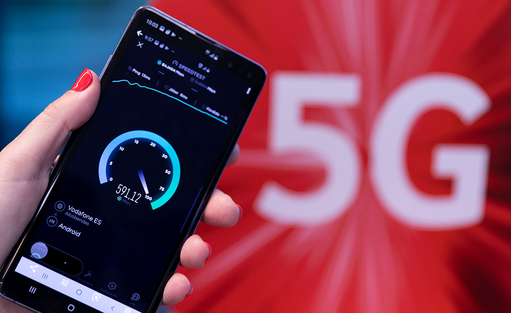 Un teléfono móvil muestra la velocidad de conexión con una red 5G y en el fondo se ve el símbolo de Vodafone y se lee 5G